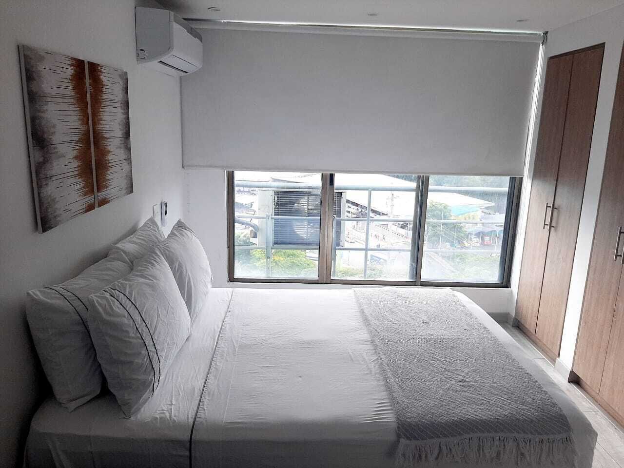 A bedroom with a large bed, a window with blinds, a wall-mounted air conditioner, and a view of buildings outside.