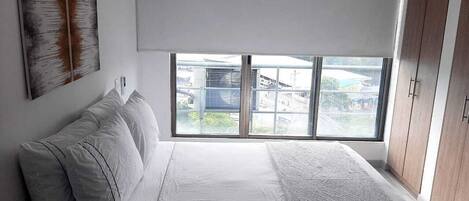 A bedroom with a large bed, a window with blinds, a wall-mounted air conditioner, and a view of buildings outside.