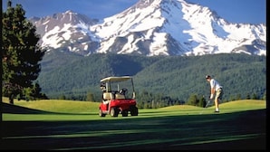 Een golfbaan met een rode golfkar en een persoon in een wit shirt die zich klaarmaakt om te putten, met besneeuwde bergen op de achtergrond.