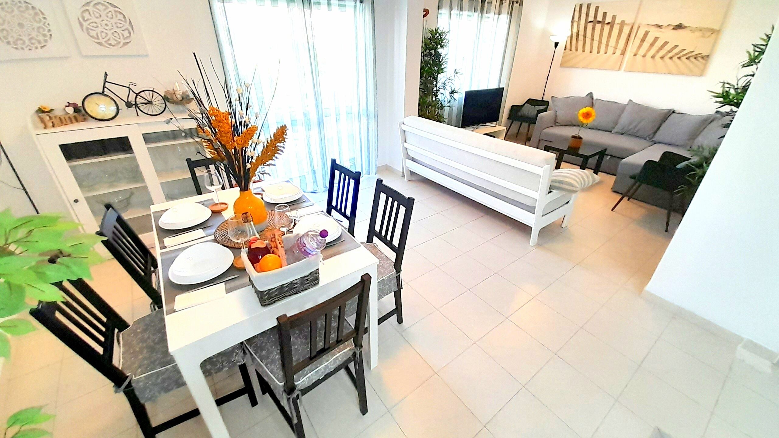 Uma sala de estar moderna com sofá, mesa de jantar posta com pratos e um vaso de flores, e uma cozinha com uma bicicleta decorativa.