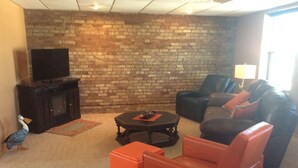 A living room with a brick wall, a television on a cabinet, a round coffee table, and a sofa with orange cushions.