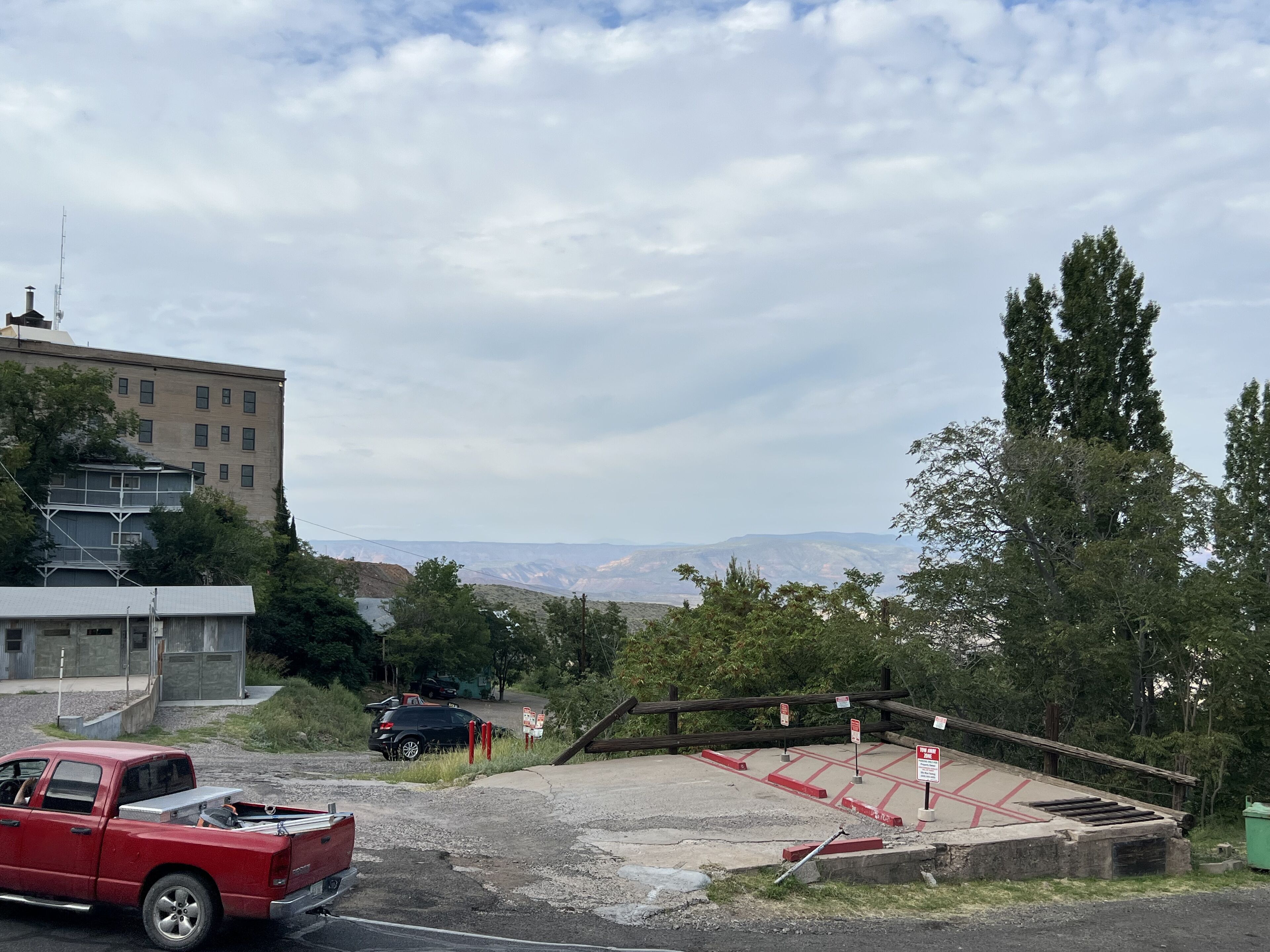 Un aparcamiento con una camioneta roja, un edificio de varios pisos y vistas a montañas lejanas bajo un cielo nublado.