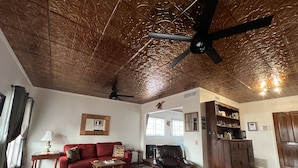 Ein Wohnzimmer mit einem roten Sofa, einem Ledersessel, einem Couchtisch aus Holz und einem gemusterten Teppich. Eine Deckenventilator, ein Kamin und ein Holzkasten mit Regalen sind ebenfalls vorhanden.