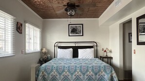Ein Schlafzimmer mit Bett, Deckenventilator, Fenster mit Jalousien, Nachttisch mit Lampe und einem gerahmten Bild an der Wand.