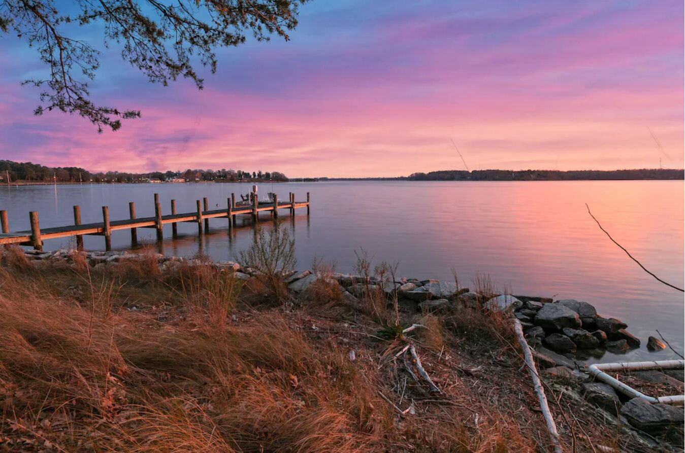 A lakeside view with a wooden dock extending into calm waters, surrounded by grassy terrain and rocks.