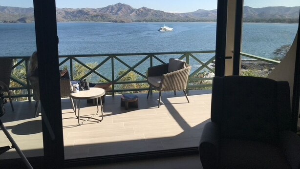 A balcony with a view of a lake and mountains, furnished with a table, chairs, and a sofa.