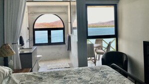 A bedroom with a bed, a desk, a chair, and a large window offering a view of the sea and mountains.