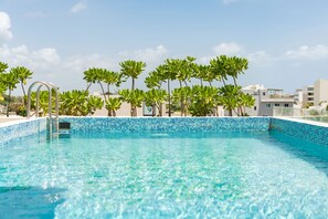 A clear blue swimming pool with a metal ladder, surrounded by palm trees and modern buildings in the background. - Boho Lofts & Studios (Playa del Carmen)