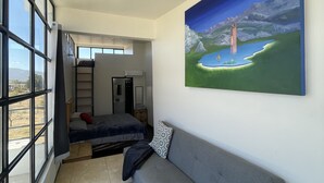 A modern room with a large window, a grey sofa, a painting of a landscape, and a bed with a red pillow. - Wine country In Valle de Guadalupe (Valle de Guadalupe)