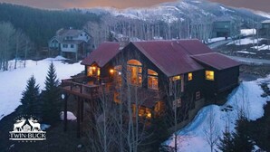 A lodge with a large deck and multiple windows, surrounded by snow-covered mountains and trees. - Hot Tub with Amazing Views- 7min to town/resort (Crested Butte)
