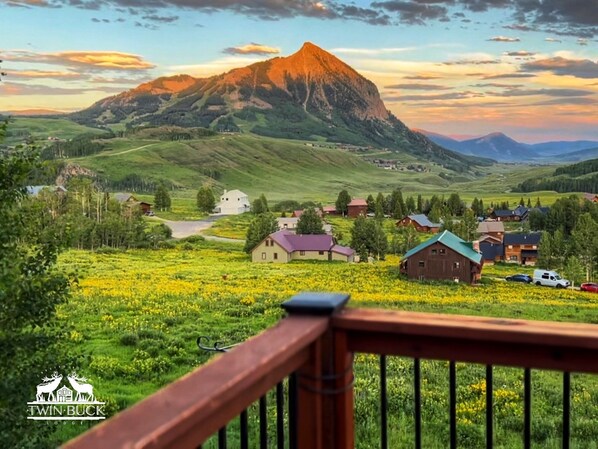 A scenic view of a mountain with a village in the foreground, featuring a wooden deck and a logo for Twin Buck Lodge.