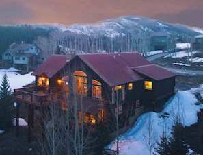 Un chalet doté d’une grande terrasse et de nombreuses fenêtres, entouré de montagnes et d’arbres couverts de neige.