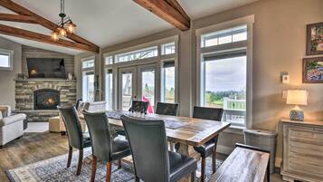 Un espace repas comprenant une table et des chaises en bois, une cheminée en pierre et de grandes fenêtres donnant sur l’extérieur.