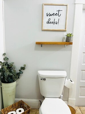 A bathroom with a toilet, a shelf with toiletries, a framed sign saying "sweet cheeks!", and a potted plant.