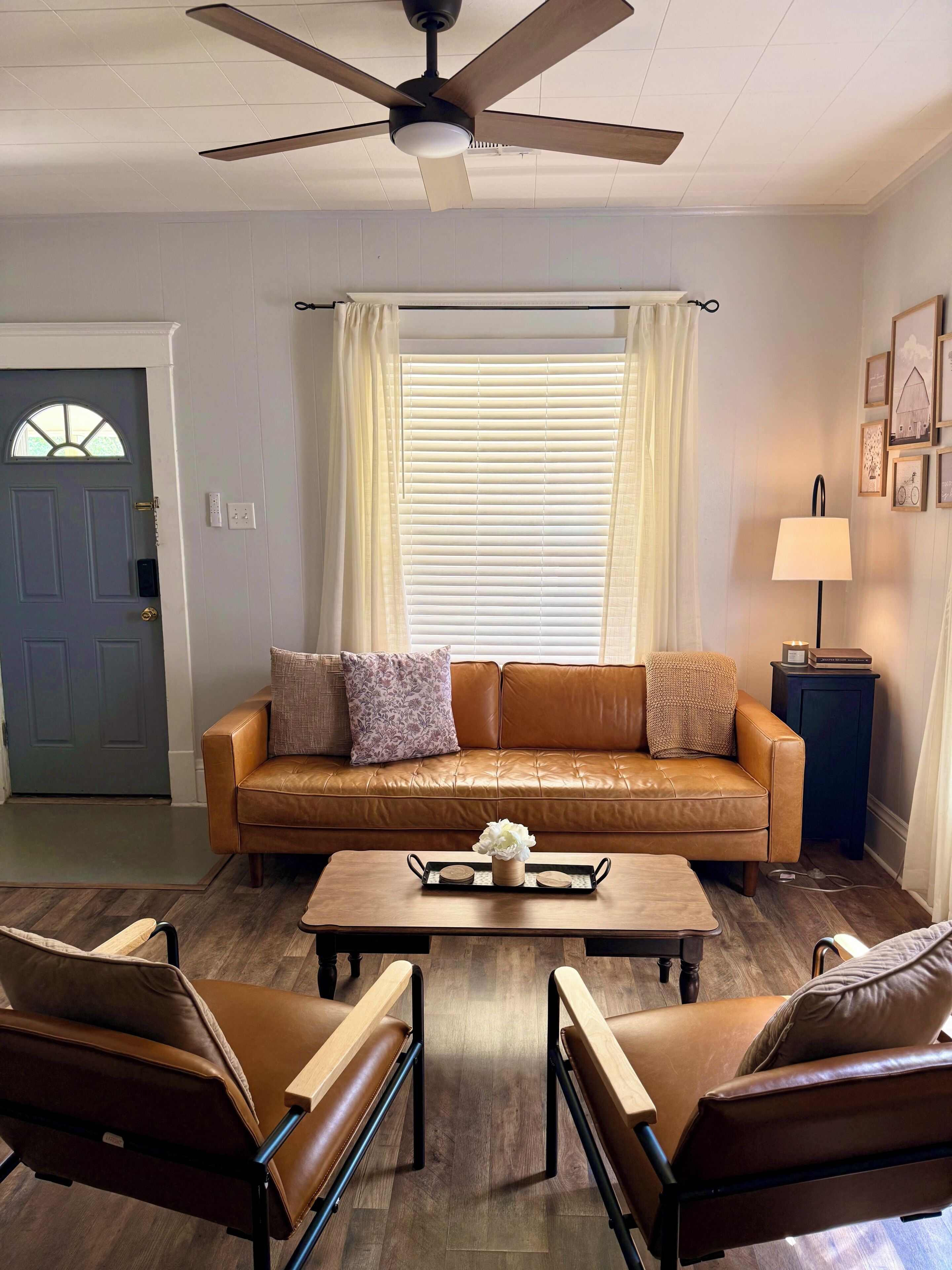 A living room with a brown leather sofa, two armchairs, a coffee table, a ceiling fan, a door, and a window with blinds.