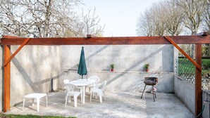 Un patio couvert avec un sol en béton, des chaises en plastique blanches, une table, un parasol vert et un barbecue.