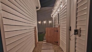 A narrow pathway leading to a wooden deck area, flanked by buildings with white siding and a metal roof. - Buckeye Bungalow charming and cozy cottage, close to everything  (Port Clinton)