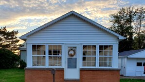 A small, single-story house with a white exterior and a brick foundation. It features a front door with a wreath, multiple windows, and a small porch with steps. - Buckeye Bungalow charming and cozy cottage, close to everything  (Port Clinton)