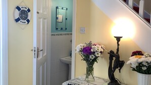 Un pasillo con una escalera, un cartel de bienvenida, un espejo, un jarrón con flores y una mesa con mantel de encaje.