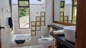 Uma casa de banho com banheira, sanita e lavatório. Tem uma janela com vista para vegetação lá fora.
