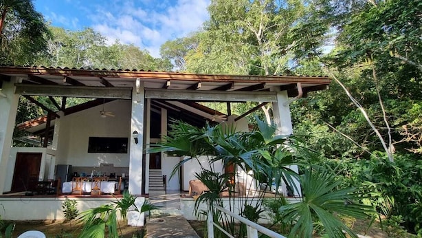 Um espaço coberto ao ar livre com área de refeições, rodeado por vegetação exuberante.