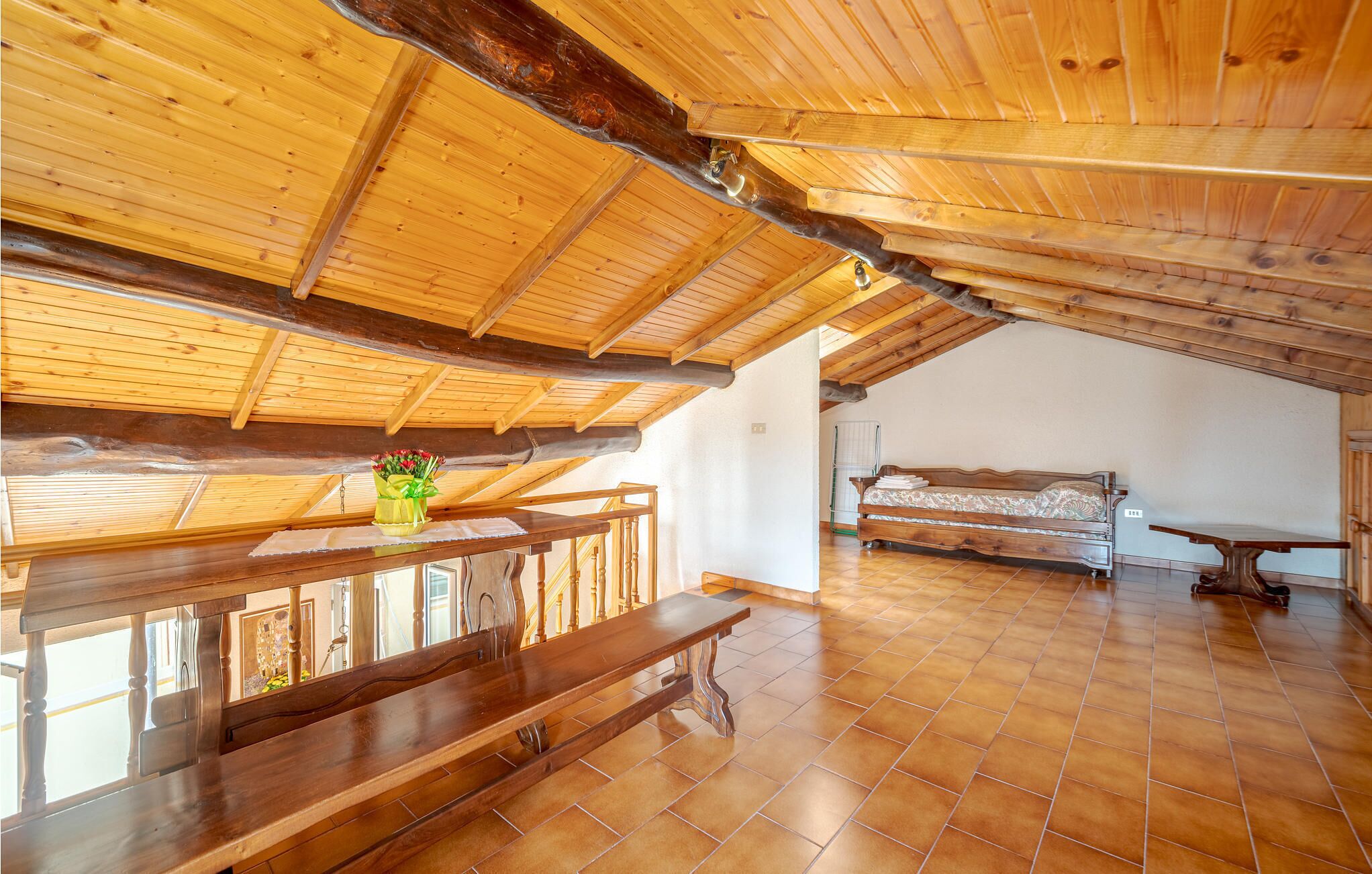 Ein geräumiges Dachzimmer mit Holzbalkendecke, einem hölzernen Bett, einer Bank und einem Tisch mit Blumen in einer Vase.