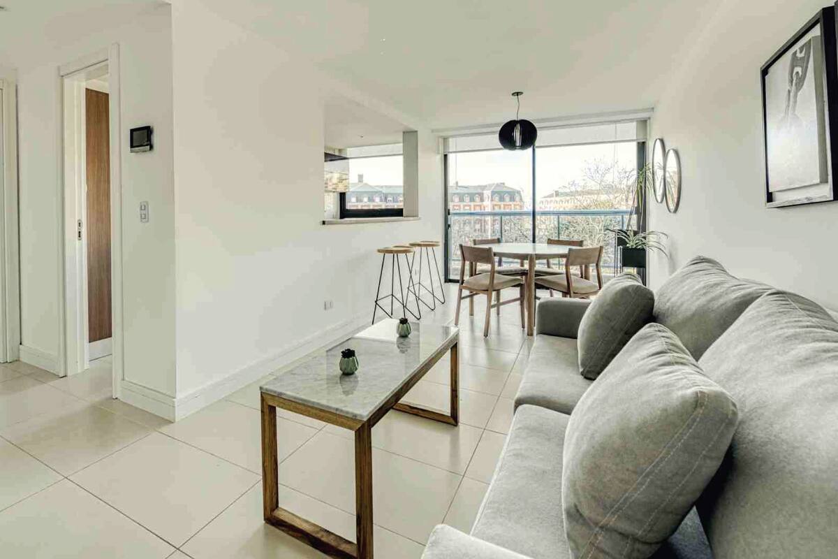 A modern living room with a grey sofa, a coffee table, and a dining area with chairs and a table. There is a balcony with a view of buildings.