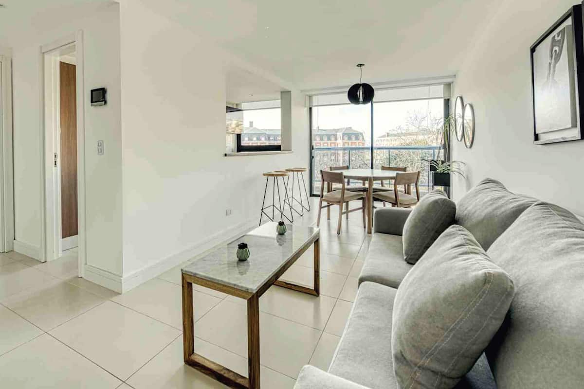 A modern living room with a grey sofa, a coffee table, and a dining area with chairs and a table. There is a balcony with a view of buildings.