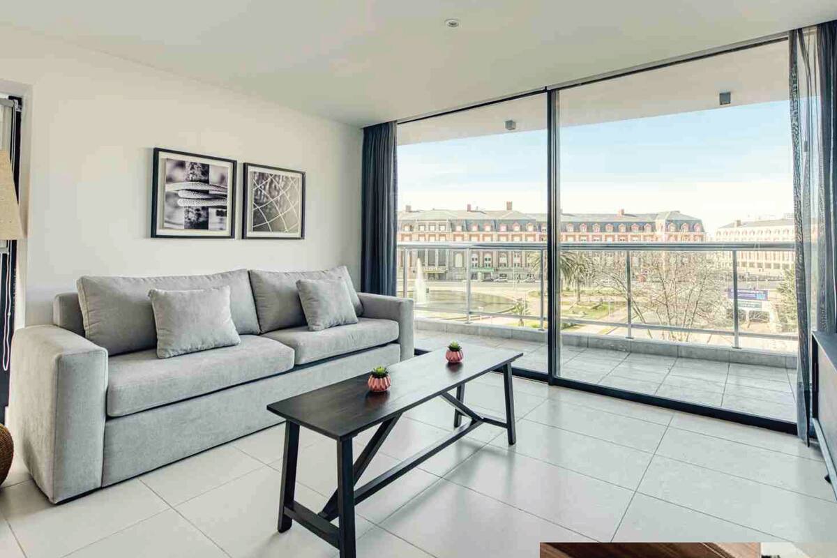 A modern living room with a grey sofa, a coffee table, and a large window offering a city view.