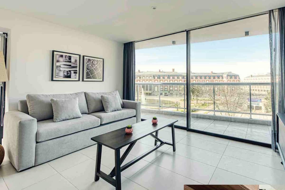 A modern living room with a grey sofa, a coffee table, and a large window offering a city view.