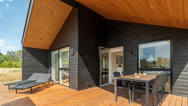 A modern house with a wooden deck, a dining area with outdoor furniture, and large windows. - Cozy home in Grenaa with kitchen (Grenaa)