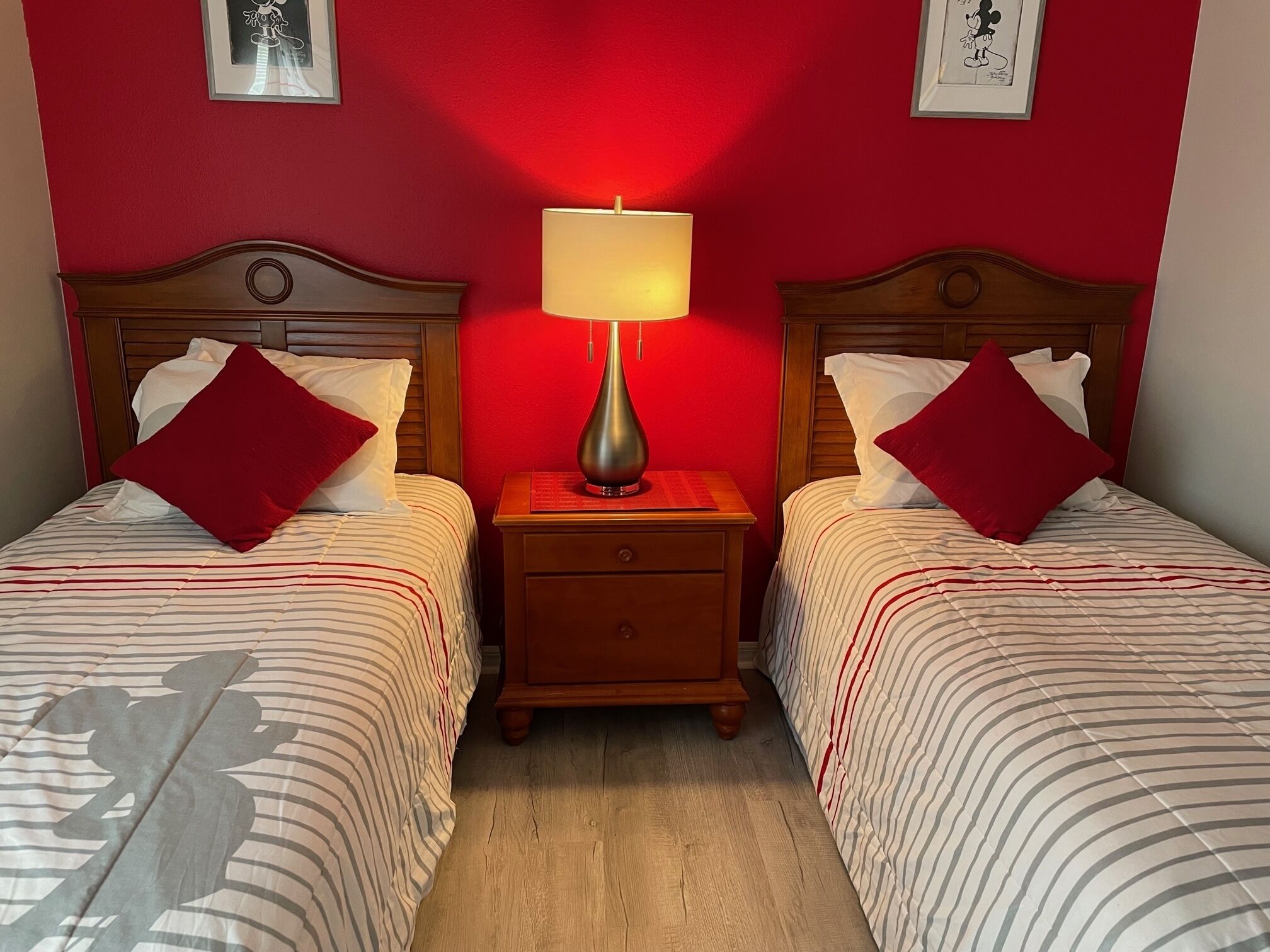 A hotel room with two beds, each with a red pillow and a striped bedspread. There is a nightstand between the beds with a lamp on it. The walls are painted red, and there are framed pictures on the wall.