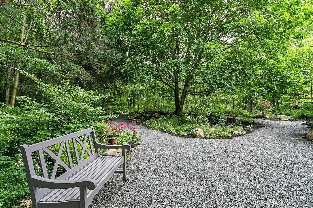 Un chemin de gravier traverse un jardin luxuriant où l’on trouve un banc en bois et des plantes en pot.