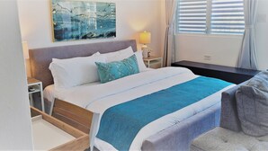 Ein ordentlich bezogenes Bett mit blauer Decke, ein Nachttisch mit Lampe und Blick ins Freie durch ein Fenster mit Jalousien.