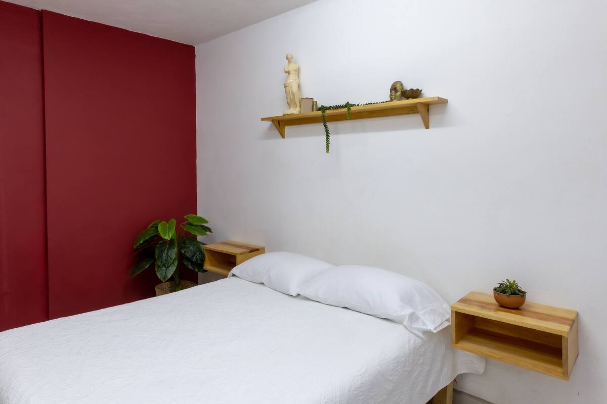 A bedroom with a bed, a red wall, a wooden shelf with decorative items, and a potted plant.