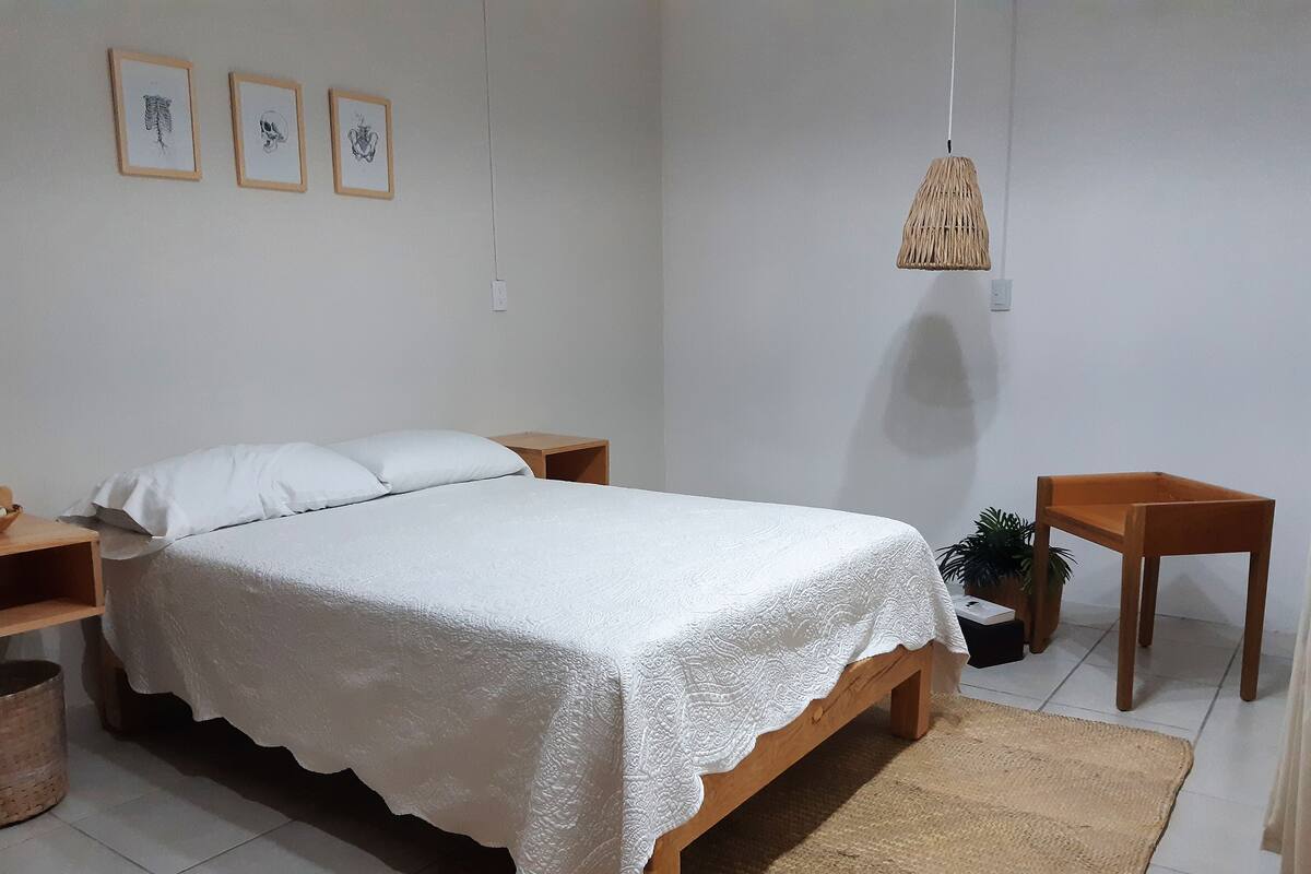 A neatly made bed with a white bedspread, a wooden nightstand, a small wooden table, and a woven rug on a tiled floor.