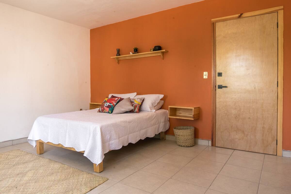 A neatly made bed with a white bedspread and colorful pillows, a wooden nightstand, a small shelf with decorative items, a wicker basket, and a wooden door.