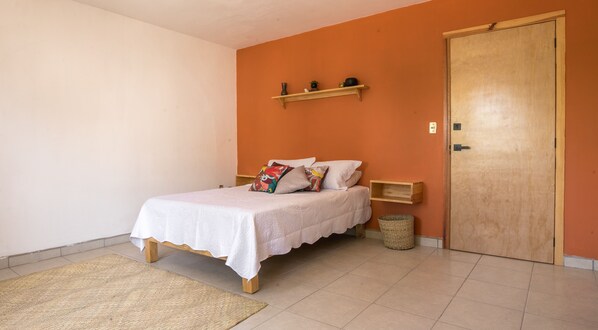 Una cama bien hecha con cubrecama blanco y almohadas coloridas, una mesita de noche de madera, un pequeño estante con objetos decorativos, una cesta de mimbre y una puerta de madera.