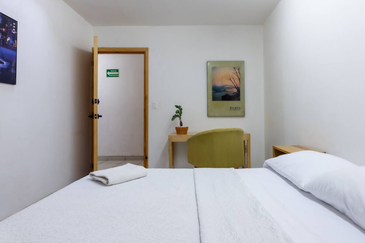 A hotel room with a bed, a chair, a desk, a potted plant, and a poster of a bridge.