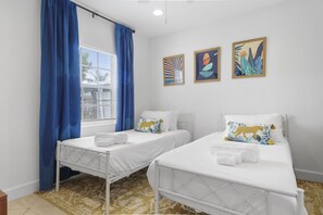 Quarto de hotel com duas camas de solteiro, uma janela com cortinas azuis e três obras de arte penduradas na parede.