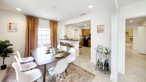 A modern dining area with a large table, white chairs, and a kitchen in the background. - Spacious Designed House -Palm Wave Stays (West Palm Beach)