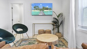 A living room with a large TV displaying a tropical beach scene, a round wooden coffee table, a green chair, and a potted plant. - Spacious Designed House -Palm Wave Stays (West Palm Beach)