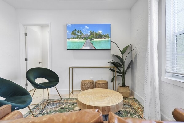 Uma sala de estar com uma televisão grande exibindo uma paisagem de praia tropical, uma mesa de centro redonda de madeira, uma poltrona verde e um vaso com planta.