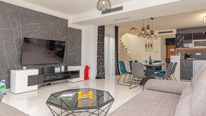 Uma sala de estar moderna com uma grande televisão de tela plana, uma mesa de centro de vidro e um espaço de refeições com cadeiras azuis.