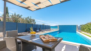 Uma área junto à piscina com móveis de vime, uma bandeja de frutas e vista para o mar.