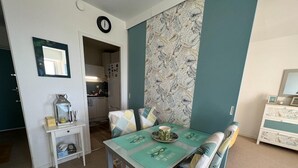 Un coin repas avec une table dressée pour quatre personnes, une cuisine en arrière-plan et une horloge au mur.