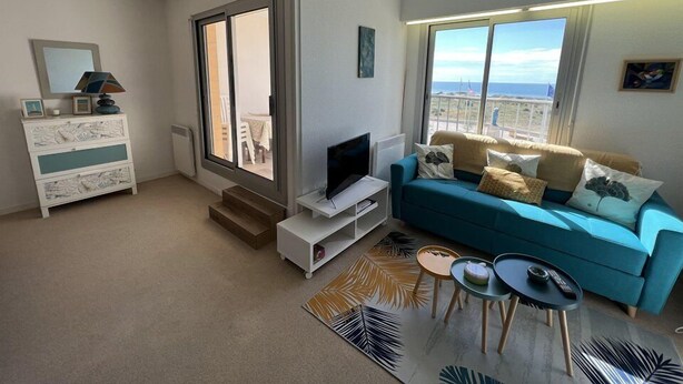 Un salon moderne avec un canapé bleu, un téléviseur à écran plat, une commode blanche et une grande fenêtre donnant sur la mer.