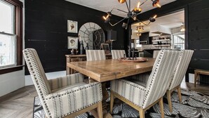 Un coin repas avec une table en bois et des chaises à motifs, un mur noir orné d’œuvres d’art, et une cuisine en arrière-plan.