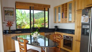 Une cuisine avec une table ronde en verre, des chaises en bois, un réfrigérateur et une fenêtre donnant sur un paysage verdoyant.