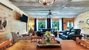 A cozy cabin interior with a wooden ceiling, a ceiling fan, a black leather sofa, a dining table with a vase of flowers, and a TV mounted on the wall. - Romantic Red River Gorge Cabin w/ Hot Tub, Pond Views & Dog Friendly (Frenchburg)